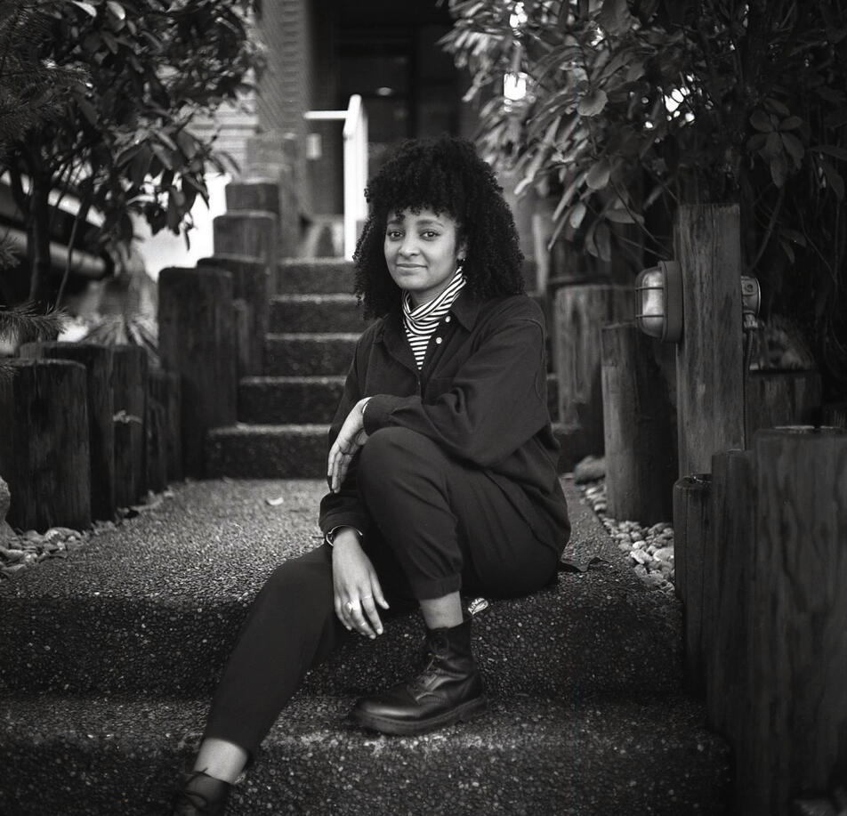 A black and white photo of Daniella, a Black woman with hair half pulled up, dressed in dark clothing, sitting on outdoor stairs.